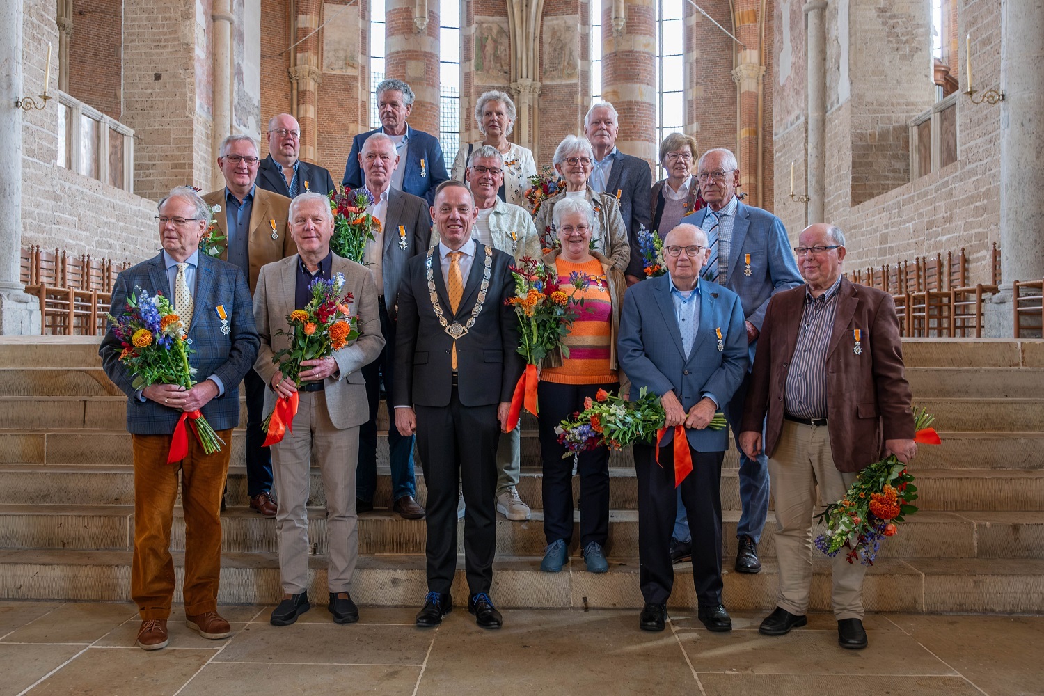 De 15 ontvangers van de Koninklijke Onderscheidingen staan samen met de burgemeester opgesteld op een stenen trap in de Lebuinuskerk. Ze hebben hun onderscheiding opgespeld.